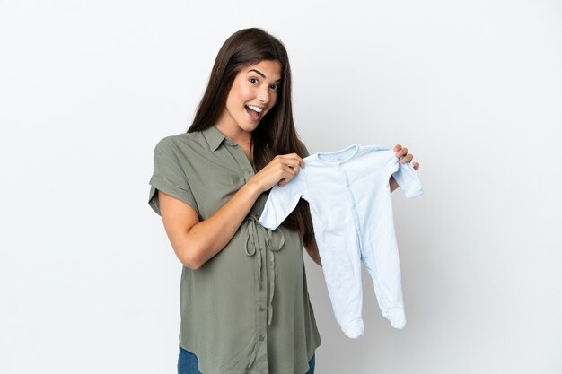 Молодая бразильская женщина изолирована на белом фоне беременной и держит детскую одежду с удивленным выражением лица
