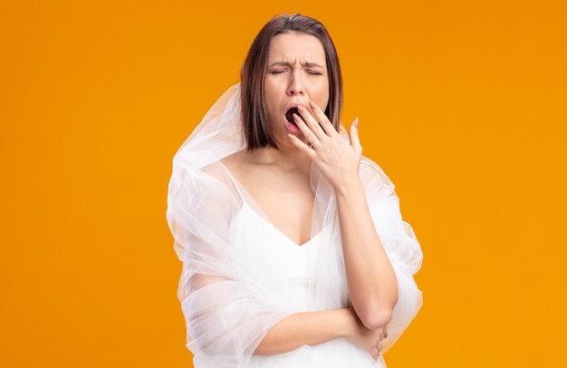 Young bride in beautiful wedding dress looking tired and bored yawning covering mouth with hand standing over orange wall