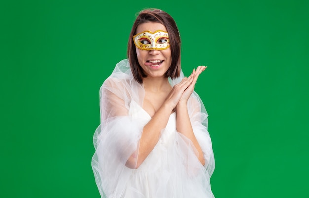 Young bride in beautiful wedding dress wearing masquerade mask looking aside happy and joyful sticking out tongue standing on green