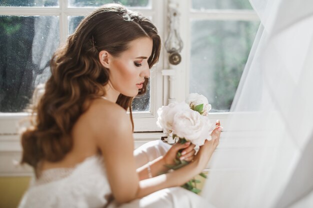 若い花嫁は、明るい朝に窓ガラスの上に座っている牡丹のブーケを保持しています