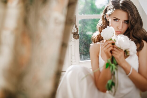 若い花嫁は、明るい朝に窓ガラスの上に座っている牡丹のブーケを保持しています