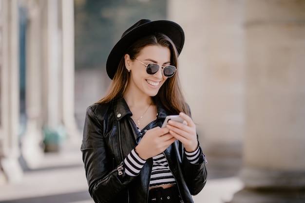 Молодая шатенка в кожаной куртке, черной шляпе на городской набережной и играет на мобильном телефоне