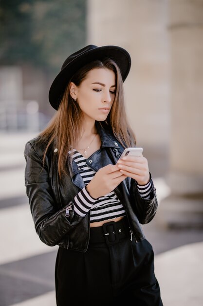 Young brown-haired girl in a leather jacket, black hat on the city promenade and plays on mobile phone