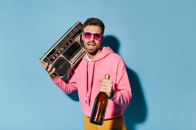 분홍색 선글라스와 후드티를 입은 젊은 브루넷 수염 남자는 고립된 파란색 배경에 레코드 플레이어와 샴페인 병을 들고 있습니다
