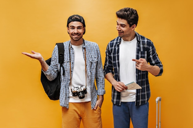 Young brunet happy men point to left at place for text on orange background Charming travelers in checkered shirts smile and hold tickets and retro camera