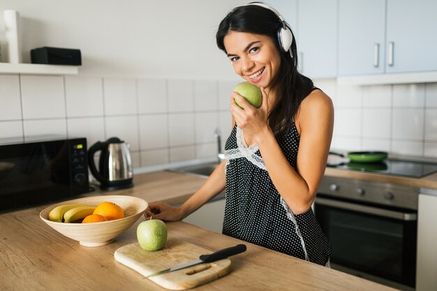 若いブルネットの魅力的な女性は、朝のキッチンで料理、青リンゴを食べる、笑顔、幸せな気分、前向きな主婦、健康的なライフスタイル、ヘッドフォンで音楽を聴く