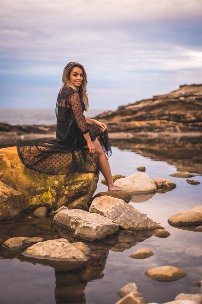 Young brunette Caucasian female in a long black transparent dress on some rocks near the sea