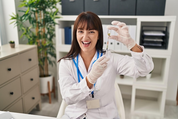 Бесплатное фото Молодая брюнетка-врач женщина, держащая шприц и вакцину, улыбается и громко смеется, потому что смешная сумасшедшая шутка.