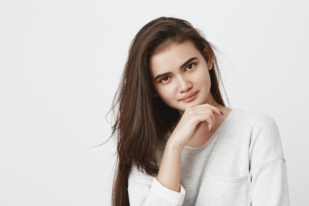 Young brunette female with oval face, dark appealing eyes having hair loose dressed in long-sleeved t-shirt holding hand under chin