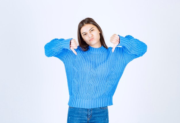 Young brunette girl in blue sweater giving thumbs down on white.