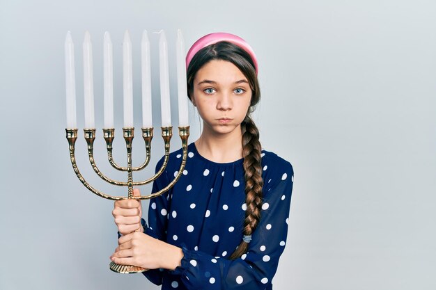 Young brunette girl holding menorah hanukkah jewish candle puffing cheeks with funny face. mouth inflated with air, catching air.