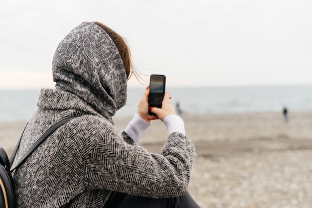 彼女​の​スマート​フォン​で​海​の​写真​を​撮って​、​曇り​の​天気​を​楽しんでいる​灰色​の​コート​と​スカーフ​の​若い​ブルネット​の​女の子