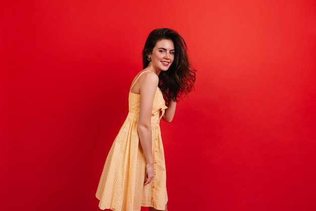 Young brunette girl is posing on red wall. Positive woman in yellow dress cute smiling.