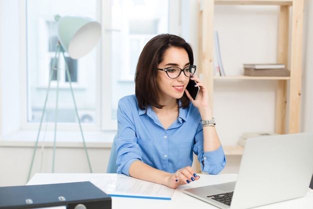 A young brunette girl is working at the table in office. She wears blue shirt and black glasses. She is speaking on phone.