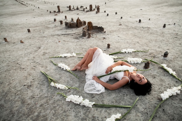 目を閉じて白いドレスの若いブルネットの少女は白いグラジオラスに囲まれた海岸に横たわっています。