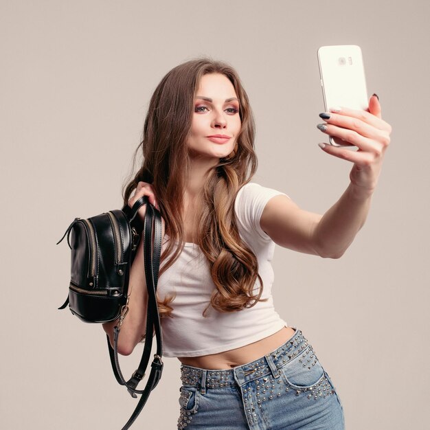 Young brunette girl with curly hair holding small backpack and smartphone Beautiful woman in casual clothes using mobile for taking selfie Pretty lady in white tshirt and jeans looking at herself