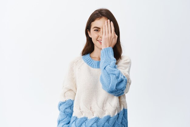 Young brunette girl with natural face and light make up, covering half of face with palm hand to show before after effect, standing against white wall