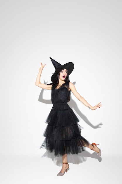 Young brunette woman in black hat and costume on white background. Attractive caucasian female model. Halloween, black friday, cyber monday, sales, autumn concept. Copyspace. Dancing, posing.