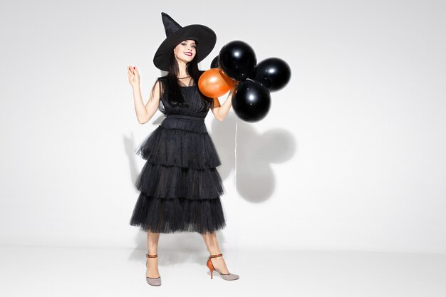 Young brunette woman in black hat and costume on white background. Attractive caucasian female model. Halloween, black friday, cyber monday, sales, autumn concept. Copyspace. Holds balloons, smiles.