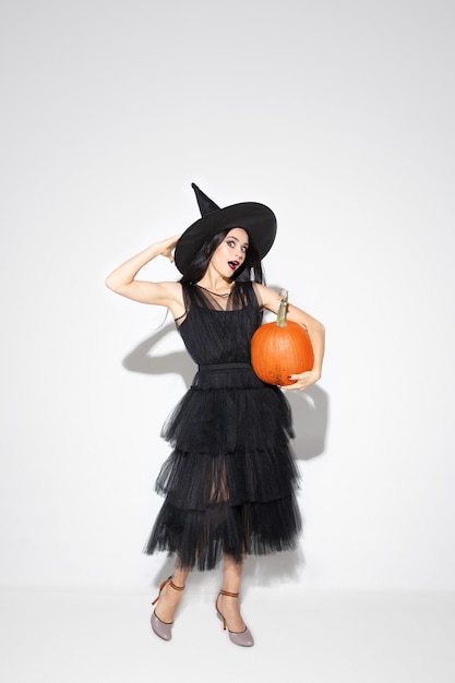 Young brunette woman in black hat and costume on white background. Attractive caucasian female model. Halloween, black friday, cyber monday, sales, autumn concept. Copyspace. Holds pumpking.