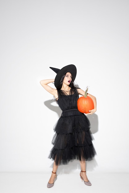Молодая женщина брюнет в черной шляпе и костюме на белой предпосылке. Привлекательная кавказская женская модель. Хэллоуин, черная пятница, киберпонедельник, распродажи, осенняя концепция. Copyspace. Держит накачку.