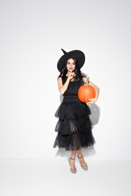 Young brunette woman in black hat and costume on white background. Attractive caucasian female model. Halloween, black friday, cyber monday, sales, autumn concept. Copyspace. Holds pumpking.