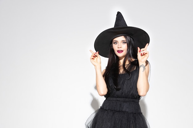Free photo young brunette woman in black hat and costume on white background. attractive caucasian female model. halloween, black friday, cyber monday, sales, autumn concept. copyspace. pointing up.