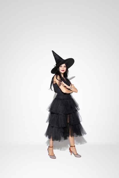 Young brunette woman in black hat and costume on white background. Attractive caucasian female model. Halloween, black friday, cyber monday, sales, autumn concept. Copyspace. Standing hands crossed.