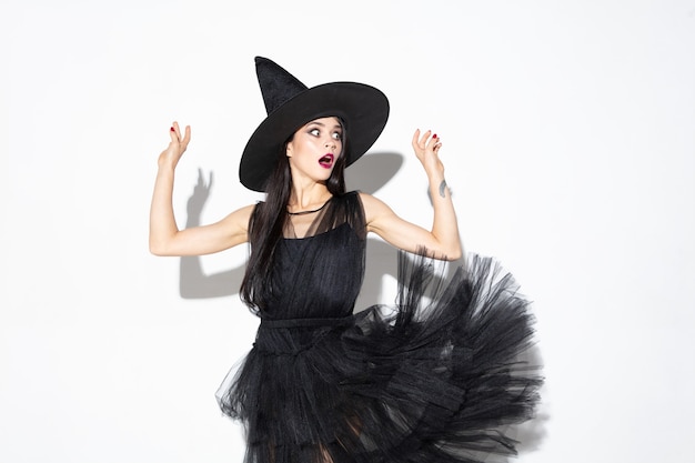 Foto gratuita giovane donna bruna in cappello nero e costume su sfondo bianco. modello femminile caucasico attraente. halloween, venerdì nero, cyber lunedì, vendite, concetto di autunno