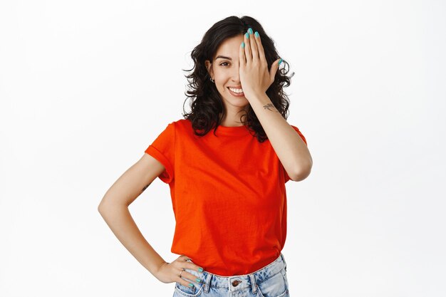 young brunette woman con one side of face, looking with pleased smile, checking vision at eyewear store, standing  on white