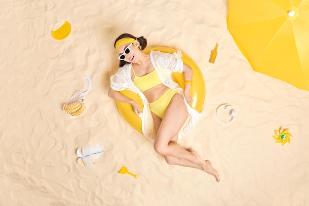 若いブルネットの女性は陽気な表情をしていますサングラスを着用し、膨らんだ水着で黄色の水着のポーズはビーチで自由な時間を過ごします熱帯の休暇中に日焼けします夏の時間のコンセプト