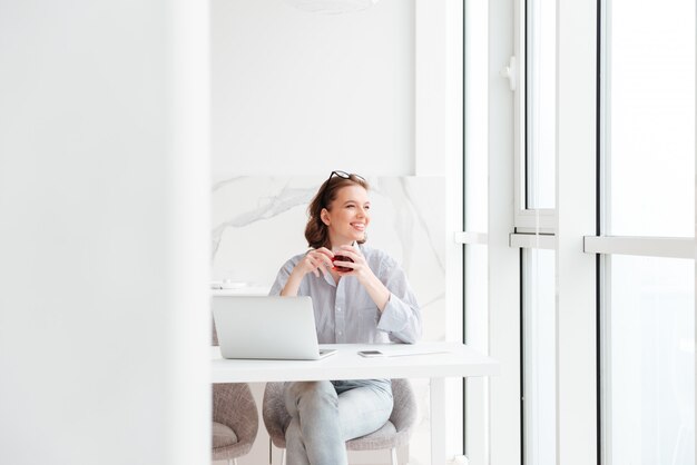Young brunette woman holding cup of tea while sitting at kitchen and looking at large window