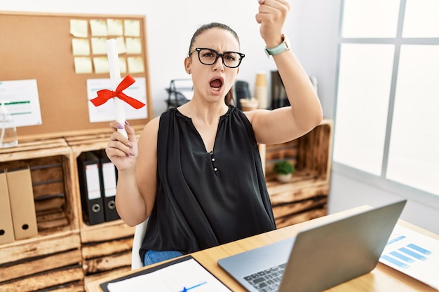 Free photo young brunette woman holding graduate degree diploma at the office annoyed and frustrated shouting with anger, yelling crazy with anger and hand raised