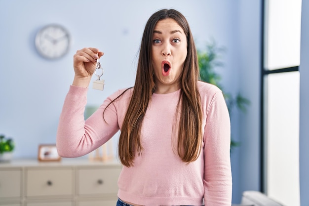 Young brunette woman holding keys of new home scared and amazed with open mouth for surprise disbelief face