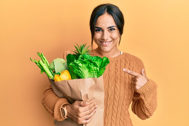 Young brunette woman holding paper bag with bread and groceries smiling happy pointing with hand and finger