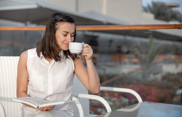 若いブルネットの女性は彼女の手で温かい飲み物と本を持って朝を楽しんでいます