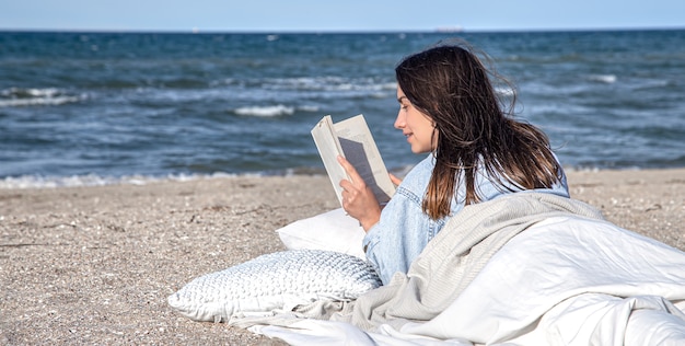 젊은 갈색 머리 여자는 담요로 덮여 해변에서 바다에 누워 책을 읽고 있습니다. 해변의 아늑한 분위기, 여름 컨셉.