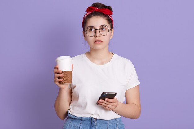 Young brunette woman isolated over lilac space holding coffee to take away and mobile while thinking about something