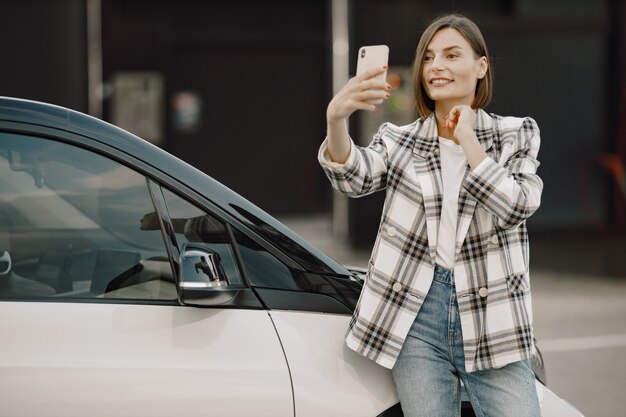 洗車場の近くで自分撮りをしている若いブルネットの女性。チェック柄のジャケットと白いTシャツを着ている女性