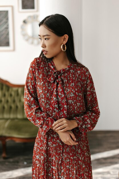 Young brunette woman in red floral dress looks into left and gently touches her arm Asian lady poses in cozy light living room