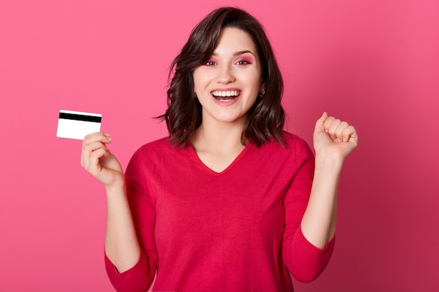 Young brunette woman shouting with happy expression and keeping fists clenched, celebrating success, holding credit card, wearing red casual shirt, standing against rosy wall.