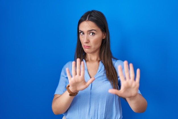 Foto gratuita giovane donna castana in piedi su sfondo blu che allontana le palme delle mani mostrando rifiuto e negazione con espressione spaventata e disgustosa fermata e proibita