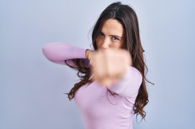 Young brunette woman standing over blue background punching fist to fight aggressive and angry attack threat and violence