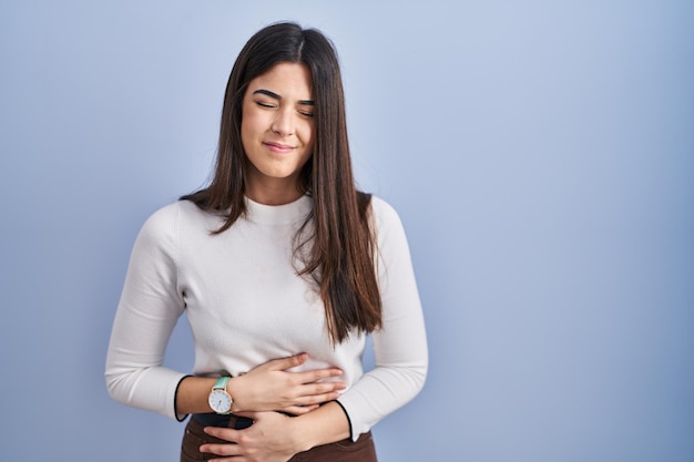 Young brunette woman standing over blue background with hand on stomach because indigestion, painful illness feeling unwell. ache concept.