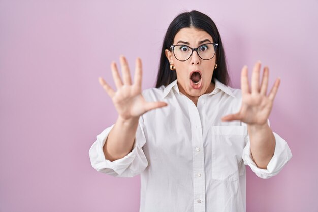 Young brunette woman standing over pink background afraid and terrified with fear expression stop gesture with hands shouting in shock panic concept