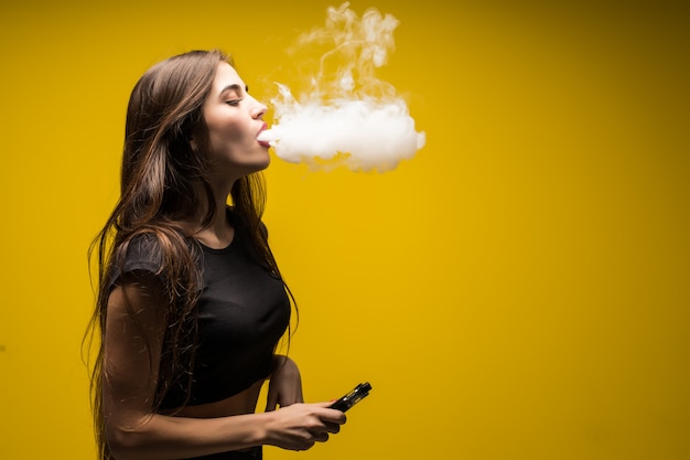 Free photo young brunette woman standing and vaping isolated on yellow wall.