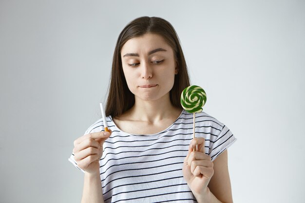 young brunette woman in striped top holding lollipop in one hand and cigarette in other, having doubtful hesitant look, feeling hard to replace bad habit with healthier one