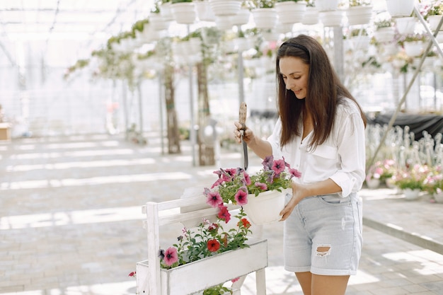 若いブルネットの女性は、庭のホースで鉢植えの植物の世話をします。白いブラウスを着ている女性