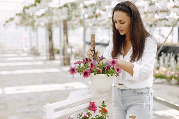 若いブルネットの女性は、庭のホースで鉢植えの植物の世話をします。白いブラウスを着ている女性