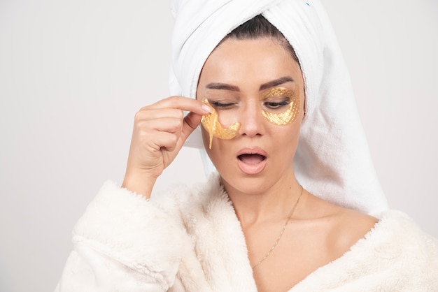 Young brunette woman wearing bathrobe applying cosmetic eye patches
